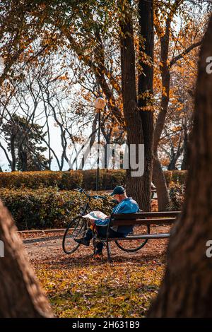 Ein Mann, der in der Herbstsaison ein Buch auf einer Bank liest. Gutes Wetter in Bulgarien. Ein Mann mit einem Fahrrad, der sich mit einem Buch entspannt. Stockfoto