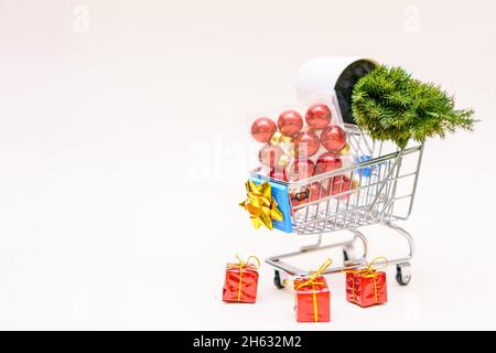 Rote weihnachtskugeln und ein künstlicher kleiner weihnachtsbaum mit Geschenken im Warenkorb auf einem hellrosa Pastellhintergrund. Weihnachts- und Neujahr Einkaufs- und Verkaufskonzept Stockfoto