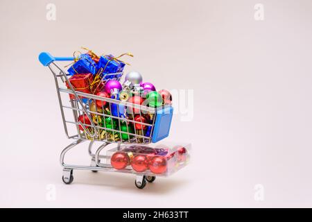 Konzept für die Weihnachtszeit Shopping Mini-Store-Warenkorb voller Geschenkboxen und Dekorationen, um den weihnachtsbaum zu schmücken, kleine bunte Kugeln auf einem hellrosa Hintergrund. Selektiver Fokus Stockfoto