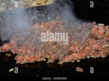 Die zerhackte Wurst wird auf dem Grill zur Vorbereitung auf die Auflaufform des Frühstücks gekocht, wobei starker Dampf aufsteigt und ein Metallspatel auf dem Hintergrund T bewegt Stockfoto