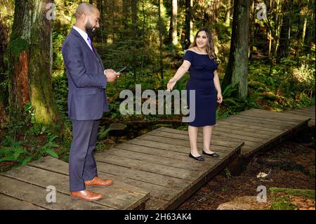 Ein junger Mann in einem Anzug und seine Verlobte, eine gut gekleidete Frau, die im Wald läuft, während der Mann anhält und etwas auf seinem Handy überprüft. Sie fragt h Stockfoto