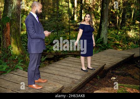 Ein junger Mann in einem Anzug und seine Verlobte, eine gut gekleidete Frau, die im Wald läuft, während der Mann anhält und etwas auf seinem Handy überprüft. Sie fragt h Stockfoto