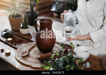 Wasser mit Kakao in den Krug gießen. Zubereitung von zeremoniellem Kakao in einem stimmungsvollen Café im Boho-Stil voller Pflanzen. Frau kocht gesundes Getränk aus Bio Stockfoto