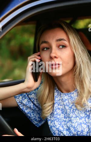Junge Frau, die auf dem Fahrersitz im Auto sitzt und mit dem Handy telefoniert. Attraktive blonde Studentin. Stockfoto