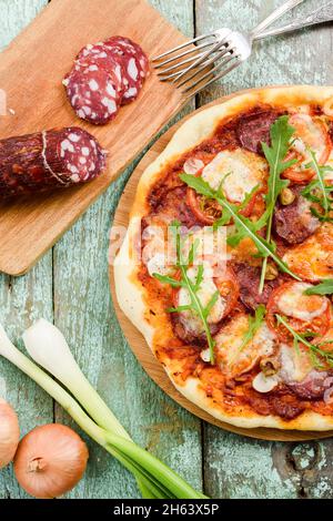 Pizza mit frischem Rucola-Salat. Salami, Zwiebel, Lauch und Gabeln auf der vertikalen Hintergrundansicht von oben Stockfoto