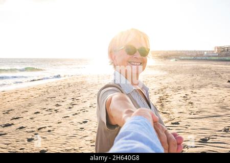 Erster Blick und pov von reifen und alten Mann hält seine Frau die Hand am Strand Spaß und genießen zusammen Sommer. Zwei glückliche Senioren im Freien mit dem Sonnenuntergang im Hintergrund. Stockfoto