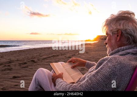 Ein reifer und alter Mann liest ein Buch, sitzt auf einem Stuhl am Strand auf dem Sand mit dem Sonnenuntergang im Hintergrund. Männliche Person genießt das Meer oder das Meer. Stockfoto