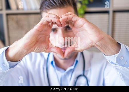 Nahaufnahme Porträt von lächelnden jungen kaukasischen männlichen Krankenschwester oder hausarzt in weißer medizinischer Uniform zeigen Herz Liebe Hand Geste. Glückliche junge Mann Arzt zeigen Unterstützung und Pflege für Patienten oder Client im Krankenhaus. Stockfoto