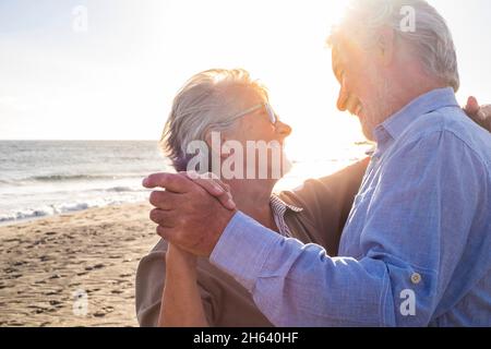Zwei alte und reife, glückliche Senioren, die im Sommer gemeinsam am Strand auf dem Sand tanzen und den Sonnenuntergang im Hintergrund genießen Stockfoto