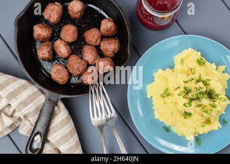Hausgemachte, unperfekte Fleischbällchen, gebraten in einer gusseisernen Pfanne, Kartoffelpüree mit frischem Lauch und Preiselbeerensause. Skandinavische Küche mit Blick von oben Stockfoto