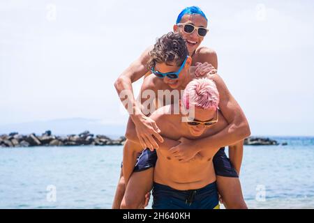 Gruppe von fröhlichen und farbigen Teenagern, die im Sommer am Strand Spaß haben und gemeinsam auf dem Sand spielen. Drei kaukasische junge Männer lächeln Stockfoto