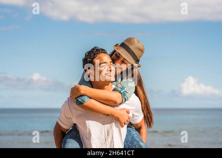 Junges Paar in der Liebe genießen Glück und Freizeitaktivitäten - Mann tragen Frau im Huckepack und lächeln mit freudig - Meerwasser und blauer Himmel im Hintergrund - Konzept der Sommerferien Stockfoto