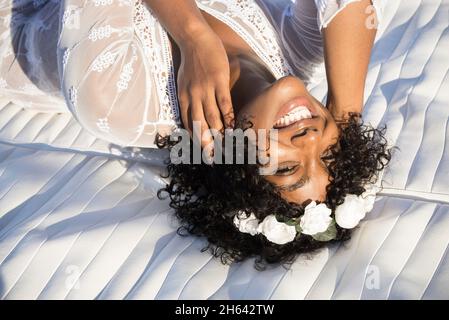 Direkt über Aufnahme einer glücklichen afroamerikanischen Frau mit lockigem Haar und floralem Haarband, die auf die Kamera schaut. Porträt einer fröhlichen schwarzen Frau in weißem Kleid. Weibliche Touristen, die Spaß im Freien haben Stockfoto