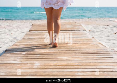 Niedriger Teil der Frau, die auf einer Holzplanke zwischen Sand in Richtung Meerwasser geht. Frau, die auf dem mit Holz beplankten Fußweg entlang des Sandstrandes zum Meer geht Stockfoto