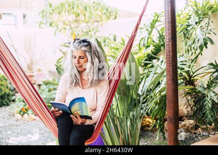 Ältere Frau liest Buch auf Hängematte auf Vorder- oder Hinterhof sitzen. Ältere Frau entspannt im Garten. Alte Frau sitzt auf Schaukelhängematte in ihrem Hausgarten und ein Buch zu lesen Stockfoto