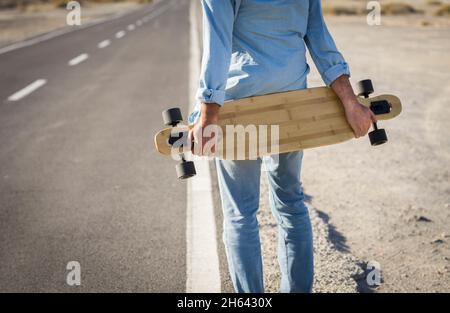 Hände von Mann hält Skateboard auf der Straße. Rückansicht des Mannes trägt Denim Kleidung hält Skateboard auf der ländlichen Straße. Sportler hält Skateboard und wartet auf der Seite der Straße Stockfoto