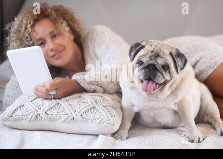 Junge Frau mit ihrem Haustier Mops mit digitalem Tablet. Netter Haustier Mops Hund mit weiblicher Besitzerin, die auf einem bequemen Bett liegt und Social-Media-Inhalte mit digitalem Tablet im Schlafzimmer zu Hause beobachtet Stockfoto