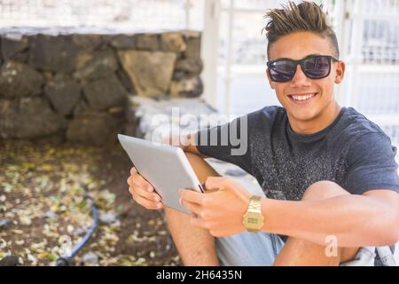 Porträt eines glücklichen Jungen im Teenageralter in Sonnenbrillen, mit einem digitalen Tablet, während er an der Grenzmauer neben dem Zaun sitzt. Lächelnder, selbstbewusster Junge, der ein digitales Tablet bediente, während er auf die Kamera schaute Stockfoto