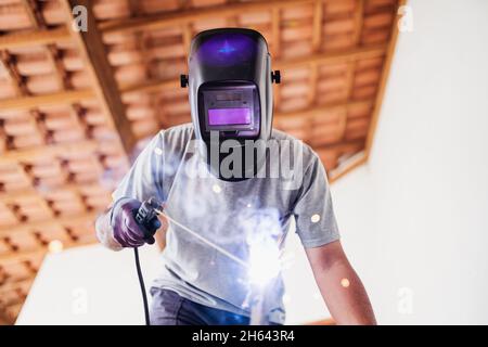 Nahaufnahme der Porträtansicht eines geschützten professionellen Maskenschweißers, der an einer Metallskulptur in einem im Bau befindlichen Haus arbeitet. Stockfoto