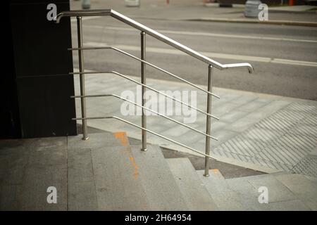 Glänzend grau gebogene Metallgeländer auf der Treppe, Ausgang aus dem Geschäft. Städtische Infrastruktur. Die Straße der Metropole im gestreuten Tageslicht. Draußen Stockfoto