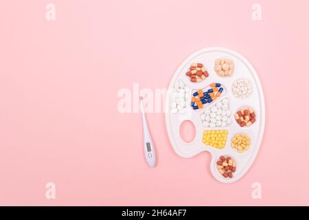 Globale Pharmaindustrie und Medizinprodukte - verschiedenfarbige Pillen, Tabletten und Kapseln auf weißer Kunstpalette mit Thermometer auf rosa Hintergrund, flach liegend Stockfoto