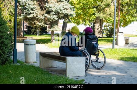 Eine ältere Frau, die sich in einem Park in Almaty um eine ältere Frau im Rollstuhl kümmert. Hilfe und Unterstützung älterer Frauen im Rollstuhl, covid-19 Lockdown Stockfoto