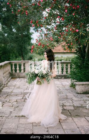 Die Braut steht unter einem blühenden Oleander mit einem Bouquet aus weißen Rosen, Pfingstrosen, Delphinien und Eukalyptuszweigen in ihren Händen Stockfoto