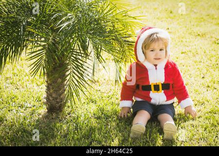 Baby santa, niedlicher Junge oder Kind in rotem Neujahr Mantel mit weißem Fell feiert weihnachten in der Nähe von grünen Palmen auf Gras Stockfoto