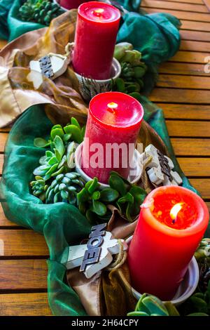 Ein Sommerweihnachtszentrum, bestehend aus drei brennenden Kerzen, umgeben von Sukkulenten, Holzdekorationen und glänzenden Bronze- und grünen Stoffen. Stockfoto