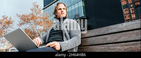 Hübscher junger Mann, der auf dem Laptop tippt, während er draußen auf einer Bank sitzt. Konzept der Work-Life-Balance. Freiberuflicher Geschäftsmann, der aus der Ferne arbeitet. Hipster Stockfoto