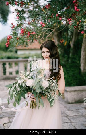 Die Braut steht unter einem blühenden Oleander mit einem Bouquet aus weißen Rosen, Pfingstrosen, Delphinien und Eukalyptuszweigen in ihren Händen Stockfoto