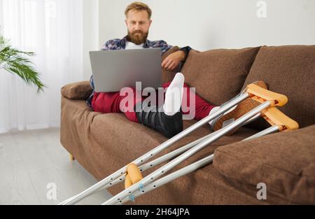 Mann mit gebrochenem Bein mit Laptop-Computer, während er zu Hause mit Krücken auf der Couch sitzt Stockfoto