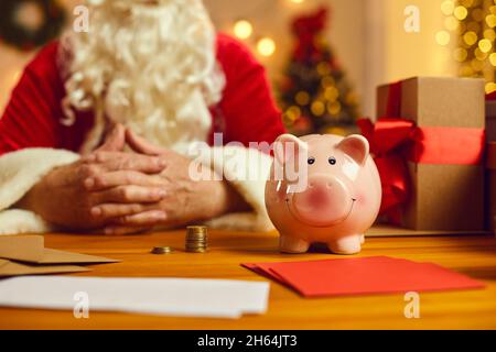 Smiley-Sparschwein auf dem Schreibtisch mit Einladungen zu Charity-Events zu Weihnachten und Weihnachtsmann im Hintergrund Stockfoto