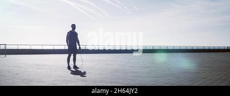 Fit jungen Mann springen mit einem Springseil im Freien. Männlicher Athlet, der morgens ein Fitnesstraining macht. Training während des Lockdown außerhalb des Fitnessraums. Stockfoto