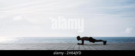 Fit junger Mann auf Plank am Meer. Fitnesstraining am Morgen im Freien. Training während des Lockdown außerhalb des Fitnessraums. Stockfoto