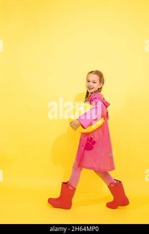 Hübsches kleines Mädchen mit zwei Zöpfen in rosa Regenmantel und Gummistiefel geht auf gelbem Hintergrund mit Ballon unter ihrem Arm. Konzept von Urlaub, Freude, Stockfoto