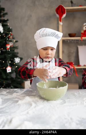 Kinder kochen zu Weihnachten. Junge Bäcker in einer Kappe und Schürze knetet Mehl für Teig für Lebkuchen oder Kekse. Prozess der Herstellung von hausgemachten Kuchen Stockfoto