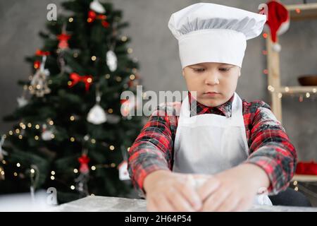 Porträt eines Bäckerjungen, der Teig zum Backen von Weihnachtsgebäck oder Lebkuchen zubereitet. Festliche Küche und Weihnachtsbaum im Hintergrund. Stockfoto