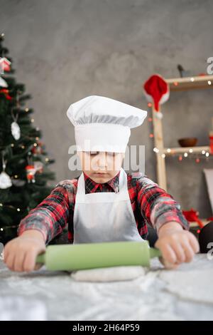 Ernsthafter Junge in der Bäckeruniform, der Kekse oder Pizza für Weihnachten zubereitet. Kind rollt rohen Teig auf den Tisch. Hausgemachtes Backkonzept Stockfoto