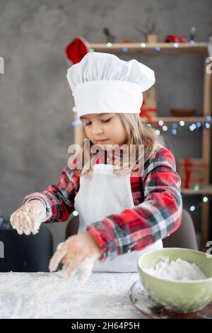 Portrait eines glücklichen Kindes, das Weihnachtskekse bäckt. Ein Mädchen in der Uniform eines Kochs knetet Teig aus weißem Mehl. Kochen hausgemachter Kuchen Stockfoto