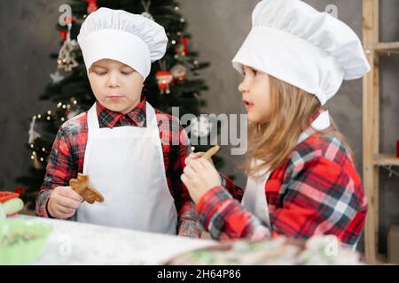 Glückliche kleine Kinder essen Weihnachtskekse in der Küche, Bruder und Schwester in Chefuniform kochen zu Hause, Kinder und WeihnachtsLebkuchen. Stockfoto