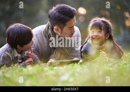 asiatischer Vater, der vorne auf Gras liegt und zwei Kindern glücklich und lächelnd Geschichten erzählt Stockfoto