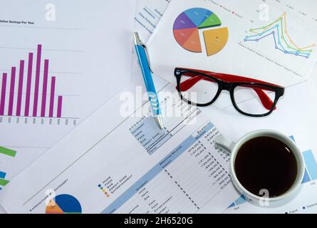 Kaffeetasse, Laptop, Notizblock und Brille über Papiere mit Zahlen und Diagrammen. Ansicht von oben Stockfoto