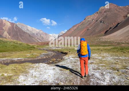 Ein Mann mit orangefarbenem Rucksack wandert an einem kalten, sonnigen Tag entlang des gefrorenen Flusses in Aconcagua vom Camp Comfluencia zum Basislager Plaza de Mulas Stockfoto