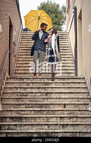 Ein junges verliebes Paar, das an einem regnerischen Tag die Treppe in der Stadt gut gelaunt hinuntersteigt. Gehen, Regen, Stadt, Beziehung Stockfoto