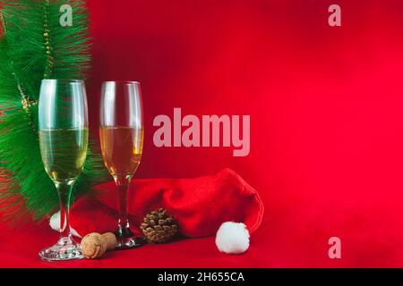Auf einem roten Hintergrund befinden sich zwei Kristallgläser mit Champagner, ein Kieferkegel, ein Korkstopfen, ein Baumzweig und ein Weihnachtsmannhut. Auf der einen Seite Stockfoto