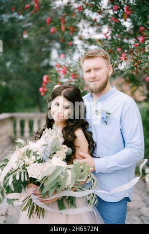 Braut und Bräutigam umarmen sich unter einem blühenden Oleander, die Braut hält einen Strauß in ihren Händen Stockfoto