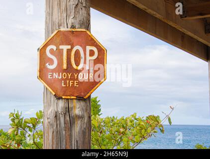 Ein abgenutztes Stoppschild mit achteckiger Aufschrift „STOP Enjoy Your Life“ ist an einem Pfosten mit dem blauen Karibischen Meer im Hintergrund angebracht. Rincon, Puerto Rico. Stockfoto