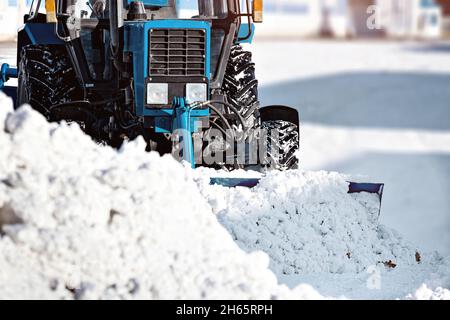 Ein blauer Traktor mit Schaufel reinigt an einem sonnigen Wintertag die Straßen von Schnee. Nach einem Schneefall löscht die Ausrüstung die Straße. Schneewehe. Stockfoto