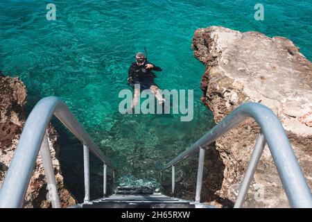 Männlicher Taucher, der in kristallklarem blauem Wasser neben der Treppe schwimmt Stockfoto